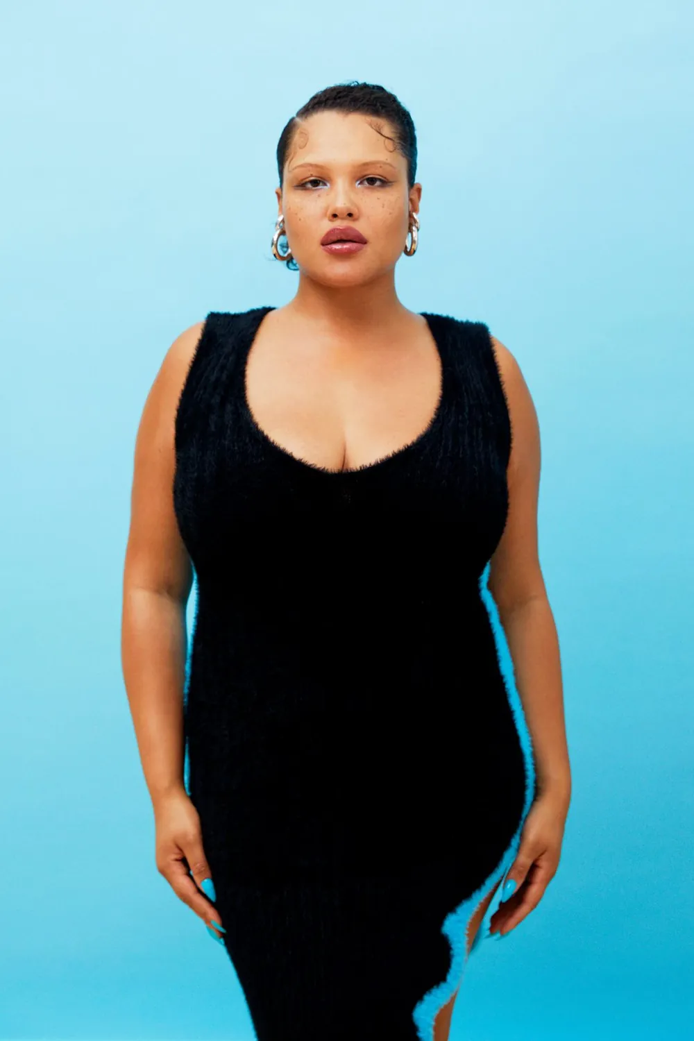 Alva Claire posing in a black sleeveless dress against a bright blue studio backdrop.