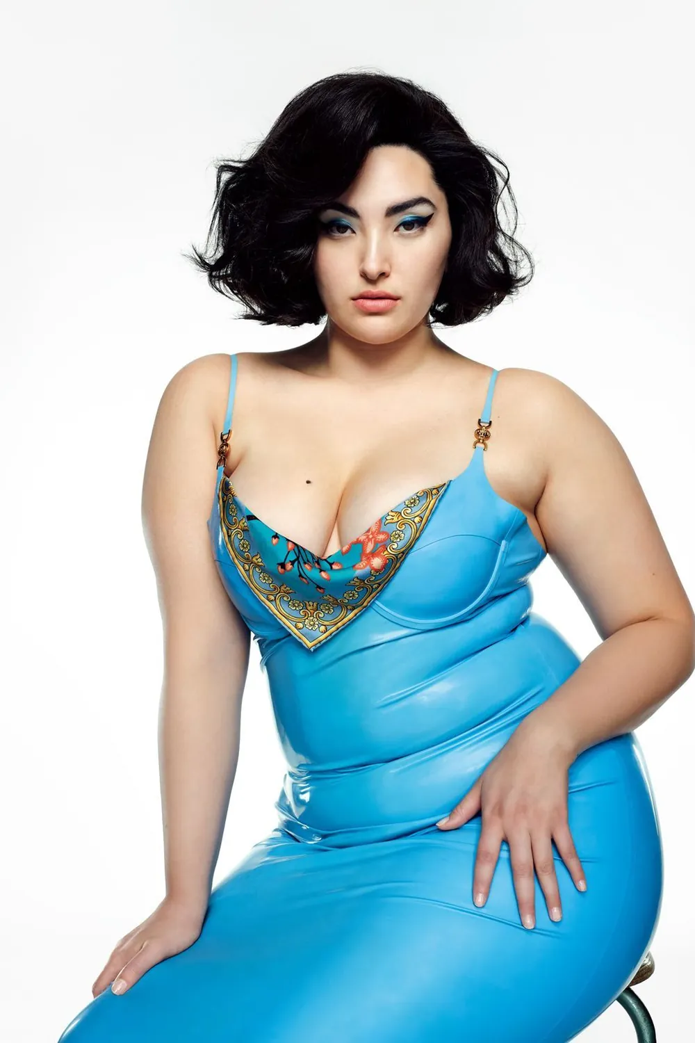 Yumi Nu posing in a blue fitted dress with a patterned neckline against a white backdrop.