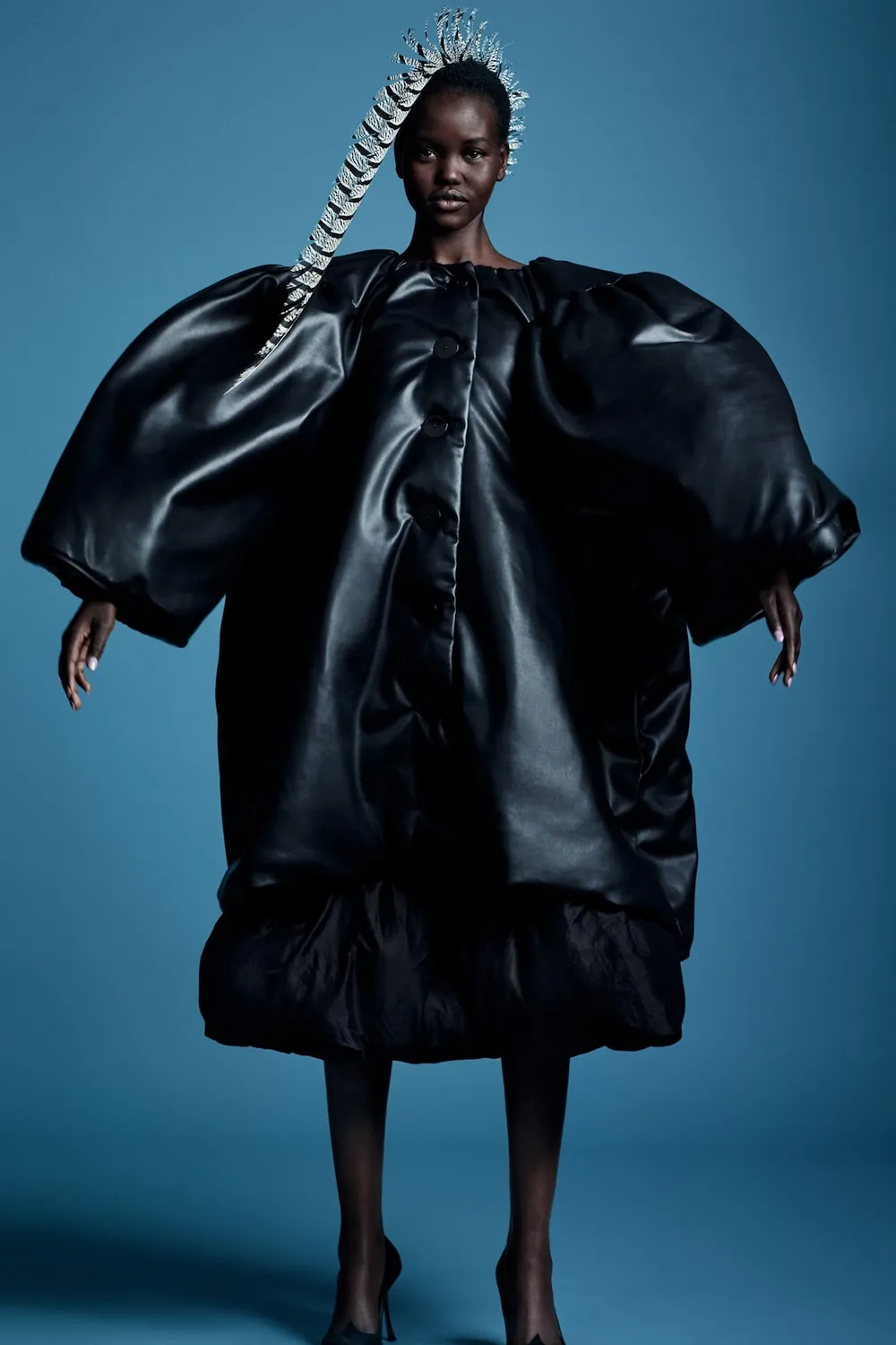 Adut Akech wearing a sculptural black padded jacket and miniskirt against a blue studio backdrop.