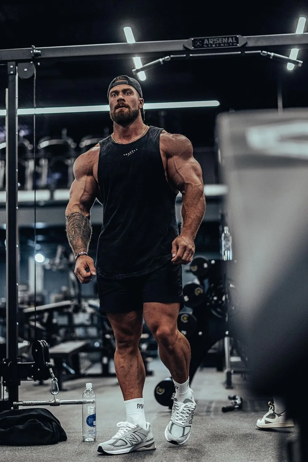 Chris Bumstead wearing a black tank top, workout shorts and a backward cap in a gym.