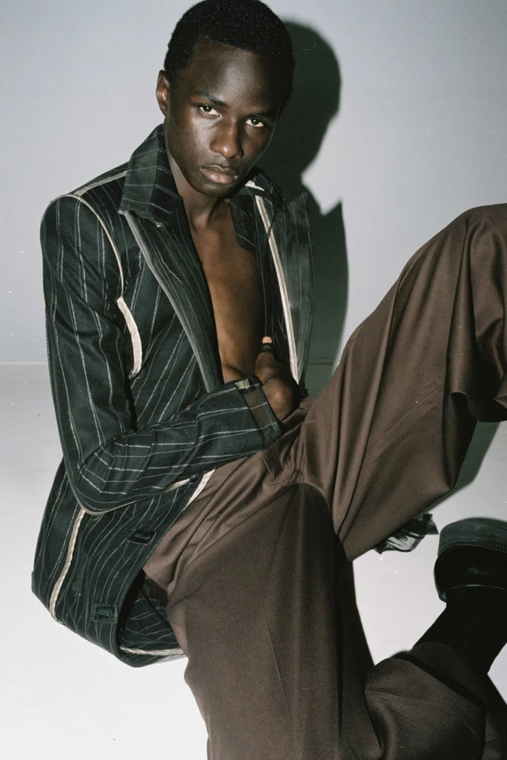 Malick Bodian seated in an open dark pinstriped blazer and loose brown trousers against a neutral studio backdrop.