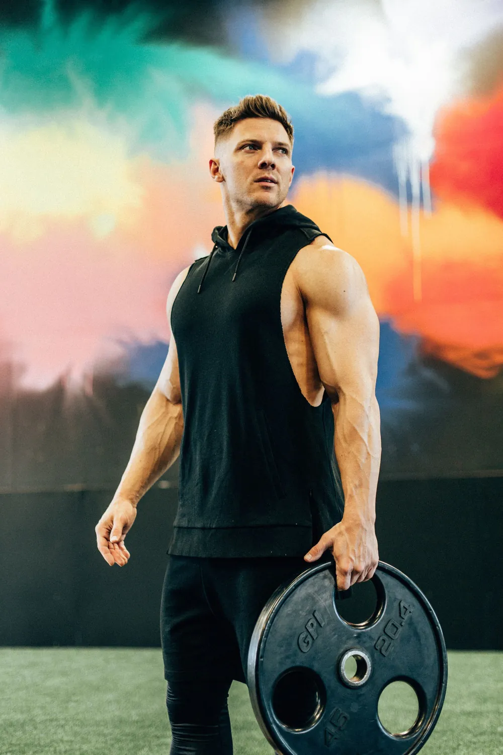 Steve Cook holding a weight plate in black sleeveless workout clothing in front of a colorful gym mural.