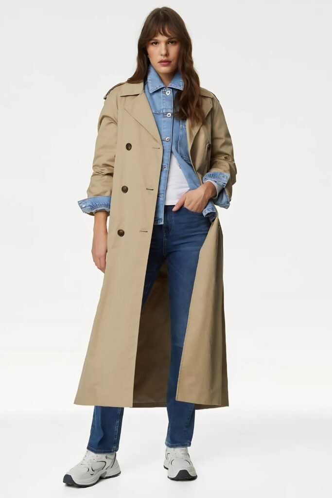 Woman in a long buff trench coat worn open over a denim shirt and jeans