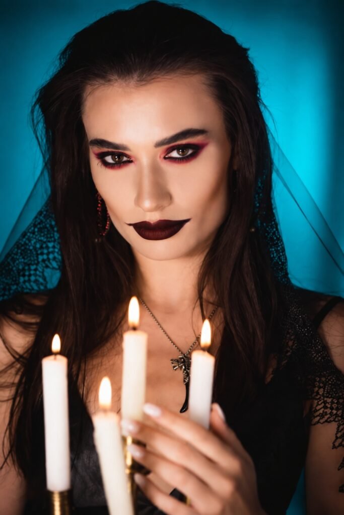 Dark berry lips and smoky eyes, holding lit candles against a teal background