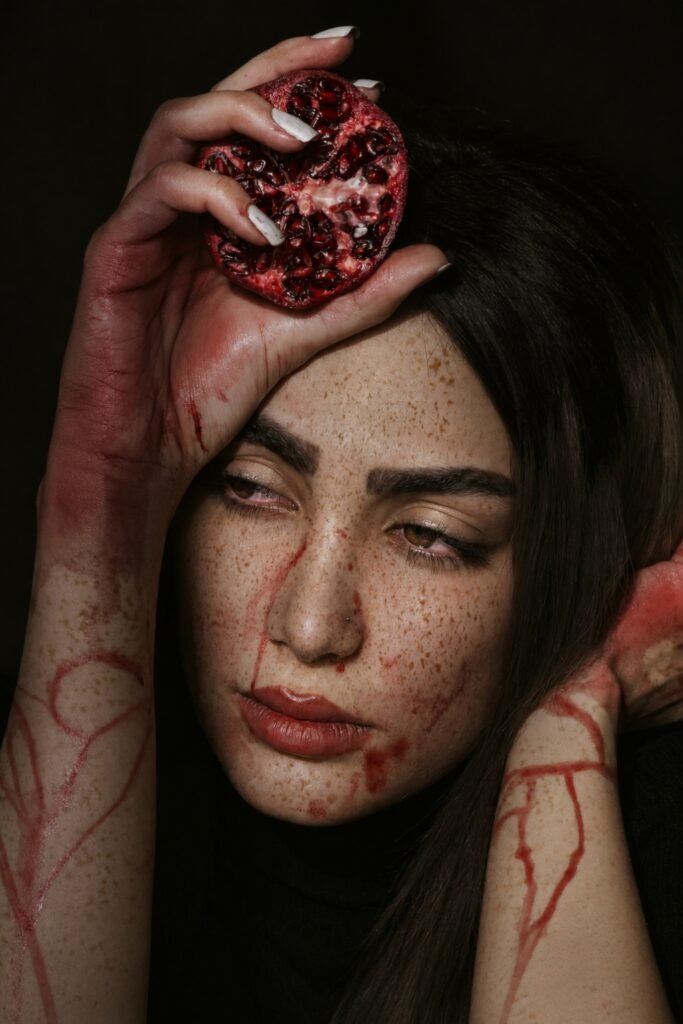Woman with dark hair holding a pomegranate, red juice stained on her hands and face
